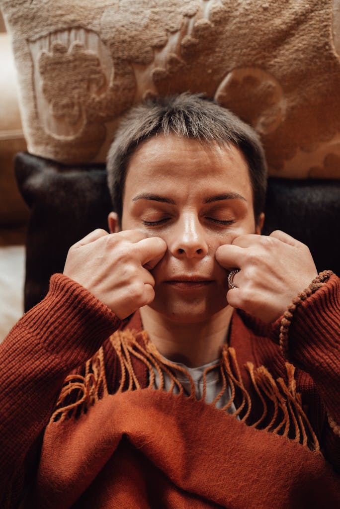 Mindful female with closed eyes and plaid massaging cheeks against decorative cushions in house room